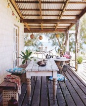 a bright tropical summer terrace with a whitewashed table, a carved wooden bench, metal chairs and textiles and lanterns attached to the roof