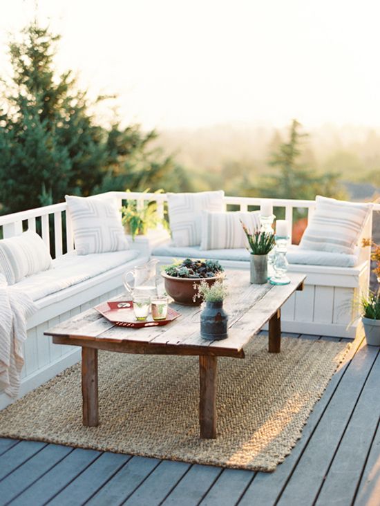 a simple and neutral summer terrace with a built-in storage bench, a wooden table, a jute rug, greenery and herbs in pots