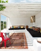 a white summer terrace with a built-in bench, dark upholstery and colorful pillows, a boho rug and a cool sea view