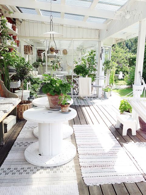 a white farmhouse terrace done with vintage rustic furniture in white, white textiles and lots of greenery in pots here and there