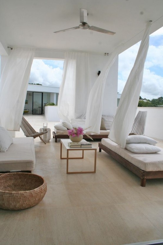 a neutral summer terrace with low wooden furniture, neutral upholstery and pillows, white curtains, candle lanterns and some blooms
