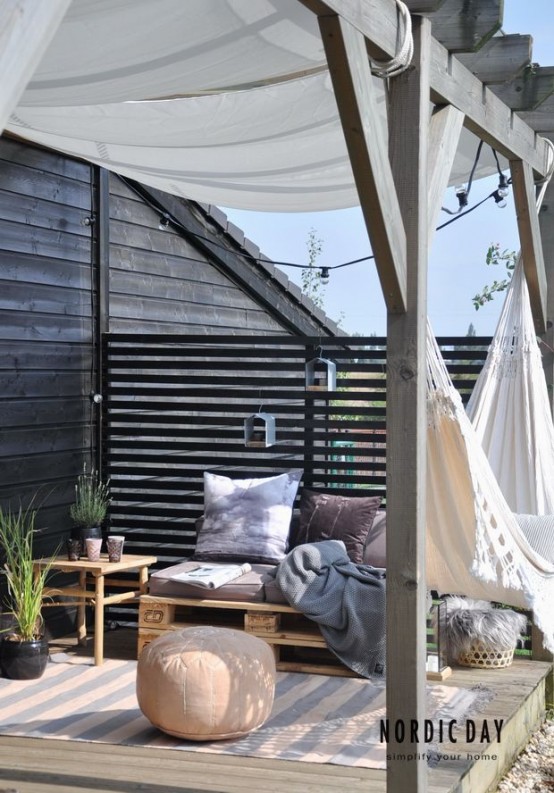 a small Nordic summer terrace with dark walls, pallet and wooden furniture, dusty textiles and a hammock plus ottomans