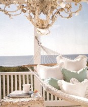 a beachy patio with a hammock and neutral and pastel bedding, with a seashell covered stool and a gorgeous seashell chandelier