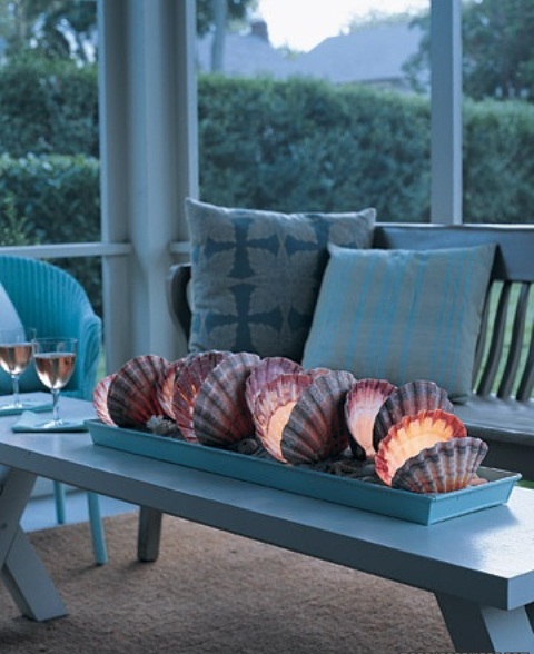 a covered beach patio with white and light blue furniture, blue printed pillows and a unique centerpiece of seashells and candles