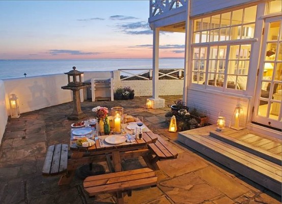 a seaside patio with simple wooden furniture, candle lanterns, blooms and a cool sea view is a very welcoming space