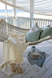 a beachy porch with a hammock, pastel and striped bedding, a stool with a candleholder of seashells and a beach view