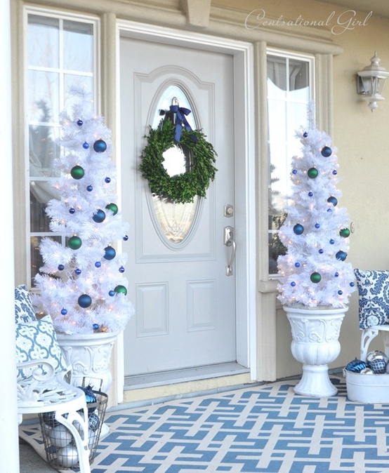 a frozen Christmas porch with white trees with navy and green ornaments, a greenery wreath and silver and navy ornaments in wire buckets