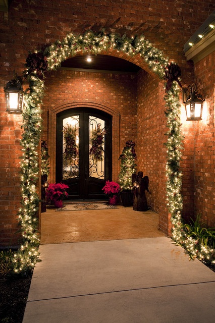 a chic and welcoming Christmas porch with a light fir garland over it, mini lit up trees and hanging plus poinsettia arrangements is very bright and cool