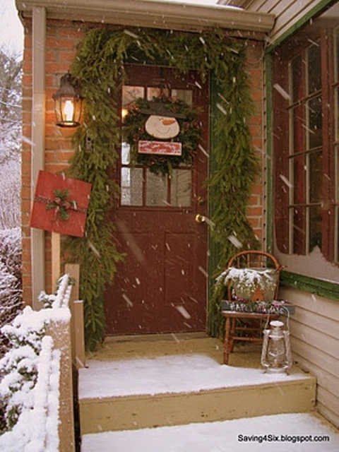 a rustic vintage Christmas porch with a fir arland framing the door, a fir wreath with a snowman, a gift box and candle lanterns