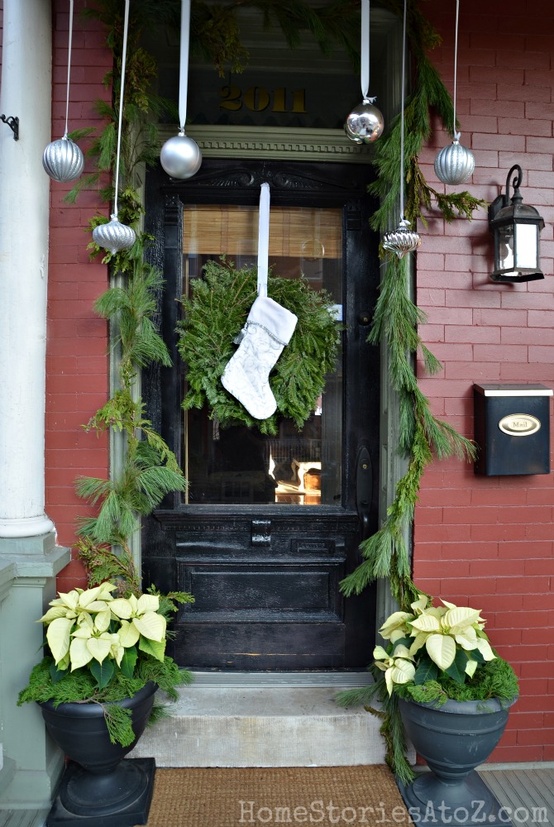 a cozy Christmas porch with silver ornaments, an evergreen garland over the door, an evergreen wreath with a stocking and poinsettias in pots
