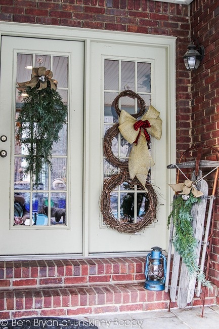 vintage rustic Christmas porch decor with a vine wreath snowman with a bow, an evergreen and burlap bow posie, a lantern and a sleigh with a burlap bow and evergreens