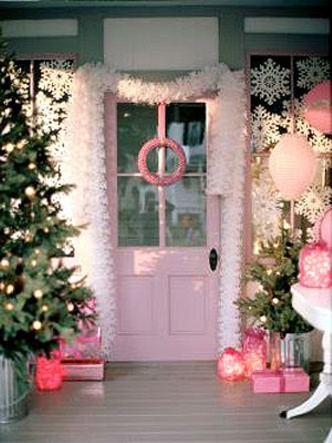a pink Christmas porch with a lit up tree, a snowy garland framing the door, a pink wreath, lanterns and baloons plus candle lanterns