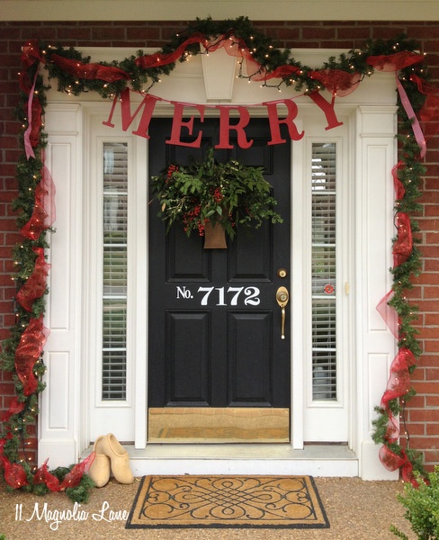 a bright Christmas porch with a lit up garland with red ribbons, a greenery arrangement on the door and a banner is lovely and bright