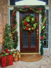 a bright Christmas porch with an evergreen and ribbon garland over the door, vine, a Christmas tree with lights and lit up Christmas gift boxes