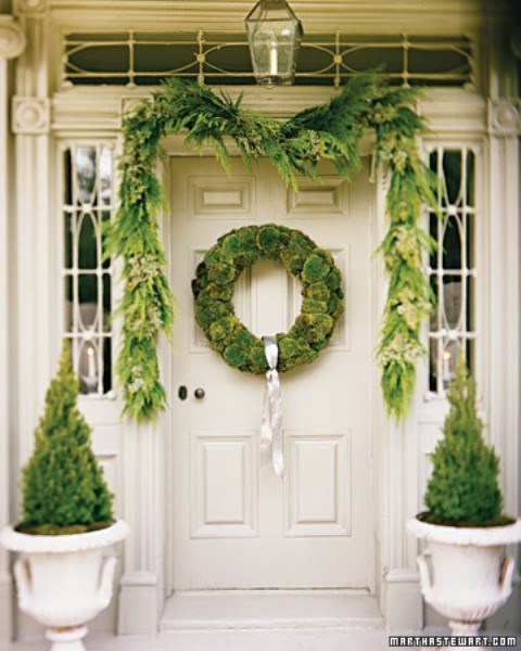 a natural green Christmas porch with a moss wreath, an evergreen garland and mini trees in pots is an out of the box idea