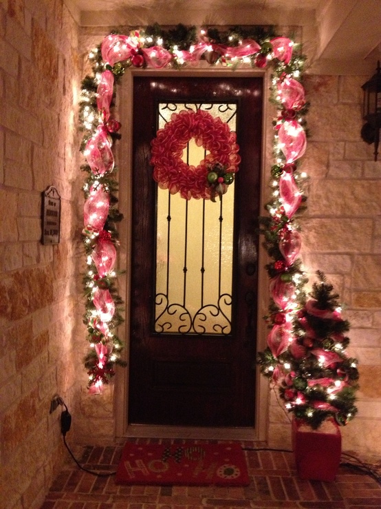 a bright Christmas porch with an evergreen and red ribbon garland framing the door, a mini tree with lights and a red wreath with green ornaments