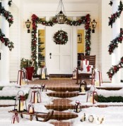 a bright Christmas porch with lots of lanterns, a sleigh, a bucket with birch branches, an evergreen garland with lots of ornaments over the door and on the pillars