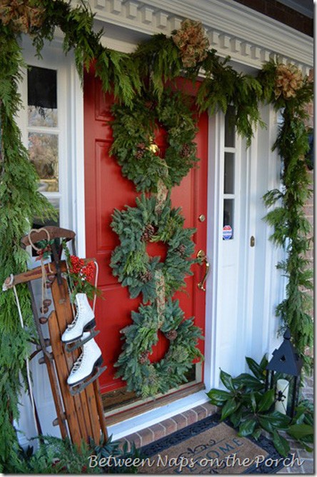 a lush rustic Christmas porch with greenery wreaths and pinecones, an evergreen garland framing the door, candle lanterns and a sleigh with bells and skates