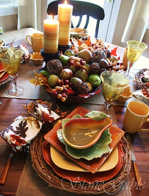 a fall tablescape in rust, orange, green and mustard, with woven placemats, layered plates, colorful glasses and mugs plus a bowl with fruits and veggies