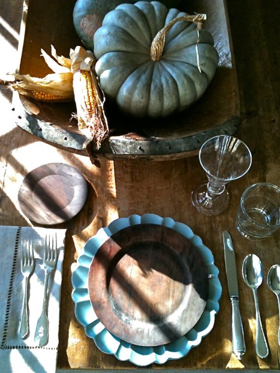 a moody rustic fall tablescape with an uncovered wooden table, shabby chc plates and a doughbowl with pumpkins and corn cobs