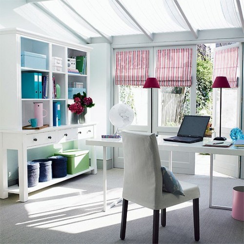 a white home office with a glazed roof, a large white bookcase, a white desk and a white chair, bold curtains and lamps and some bright boxes