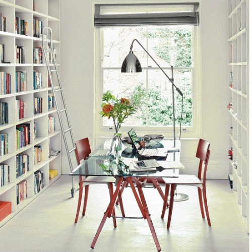 a large white home office with large bookcases, a glass top shared desk, red chairs, a ladder and a table lamp