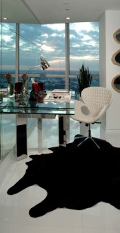 a small and bold white home office with a glass wall and a city view, a mirror and glass desk, a white chair, a black rug and a metal table lamp is a lovely and bold space for working