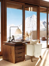 a chalet home office with a glazed wall and a mountain view, a wooden desk, a creamy chair, a table lamp and some branches in a vase