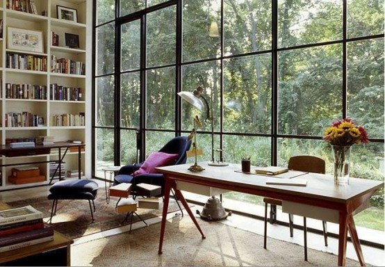 an inviting home office with a glazed wall and a forest view, a built-in bookcase, a wooden desk, some lovely chairs and tables is a lovely space to be in