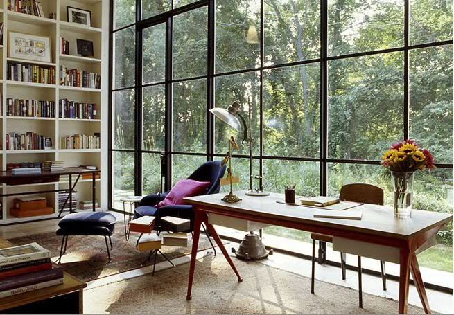 an inviting home office with a glazed wall and a forest view, a built in bookcase, a wooden desk, some lovely chairs and tables is a lovely space to be in