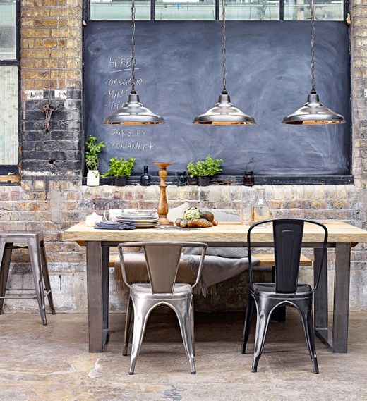an industrial dining room with brick walls, a large chalkboard, an industrial table with soild black legs and metal chairs