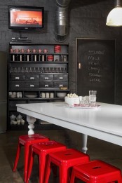 a dark and moody industrial dinign space with black walls, a black storage unit, exposed pipes, a white vintage table and hot red metal chairs
