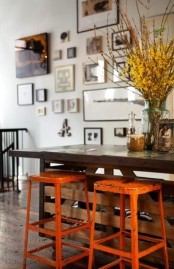 an industrial dining space with a dakr table, orange metal stools and a gallery wall, bloomign branches in a vase