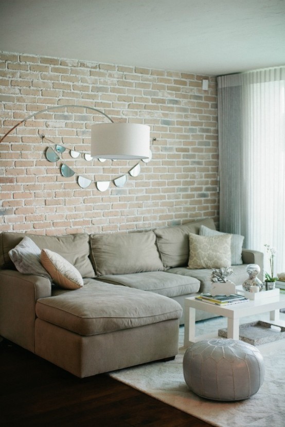 a neutral living room with muted brick walls that bring texture and interest to the space, with shiny touches nd a leather ottoman