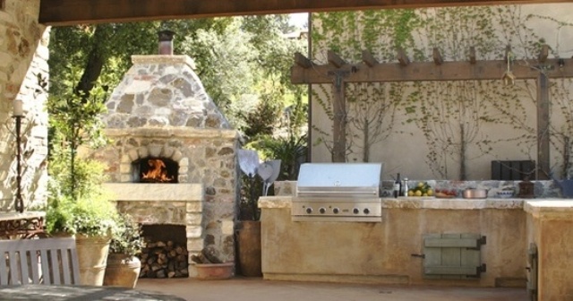 a traditional outdoor kitchen of stone with a grill and a pizza oven plus greenery around