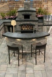 a dark bbq area of stone and bricks, with black countertops, a grill and a pizza oven plus a dining space