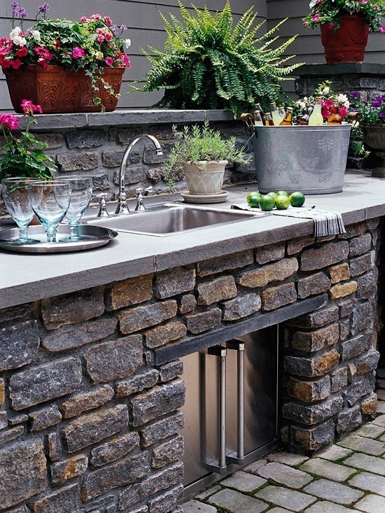a traditional kitchen with stone, bricks and a storage unit with metal doors, a sink and potted greenery and blooms