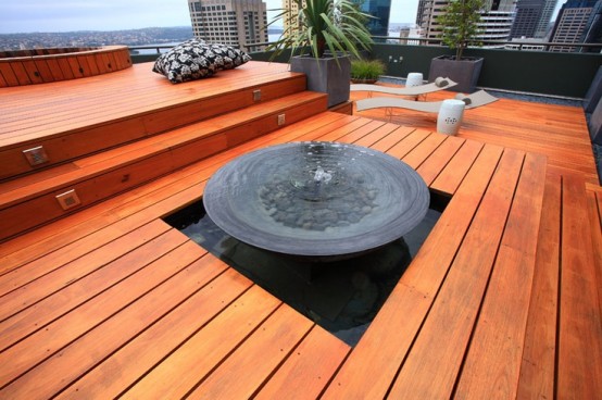 a minimalist deck done with orange wood, pillows, loungers, a fountain and potted greenery