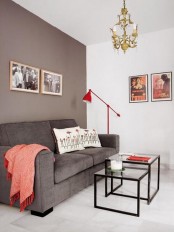 a modern and laconic living room with a grey accent wall, a grey sofa, a red blanket and a red floor lamp, coffee tables and a gold chandelier