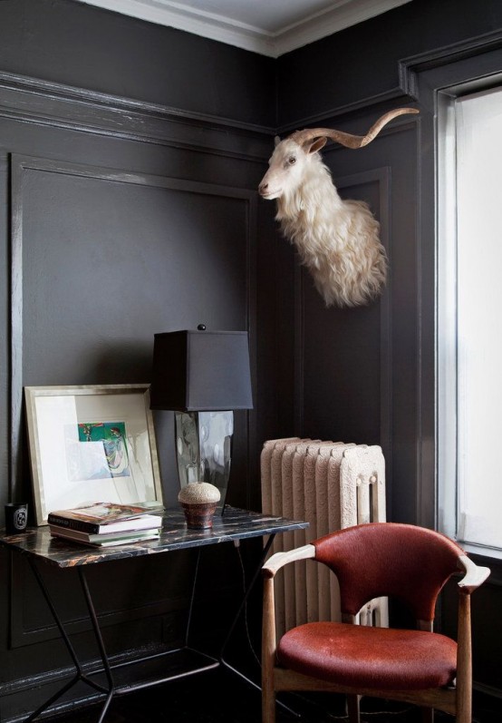 a whimsical and moody nook with black walls, a chic table, artwork and an elegant lamp, a deep red leather chair and taxidermy on the wall