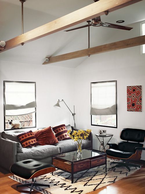 a beautiful mid century modern living room with a grey sofa, black chairs, a chic coffee table, a printed rug and wooden beams, red pillows and an artwork