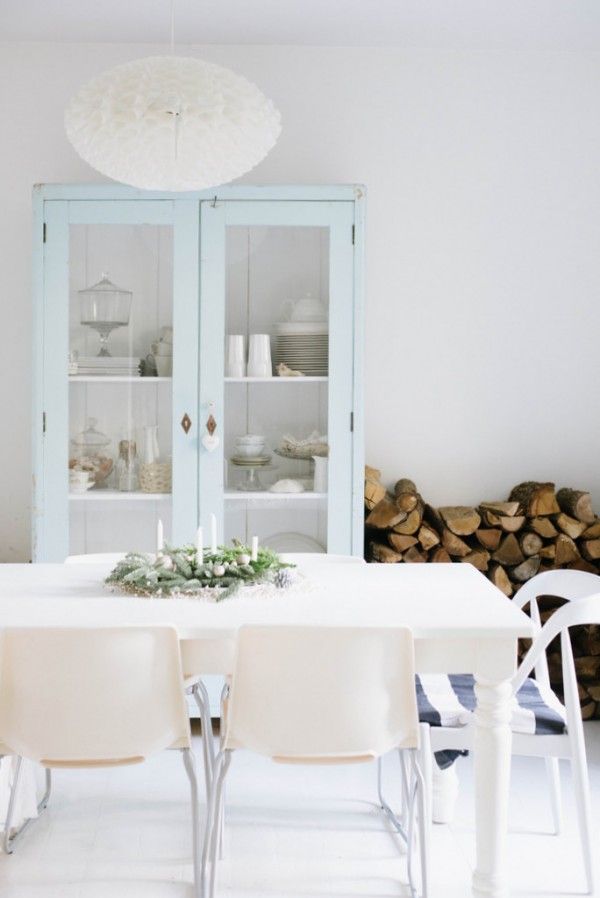 a welcoming Nordic dining space with a light blue vintage buffet with glass doors, a white vintage table and mismatching white chairs plus firewood for a cozy feel here