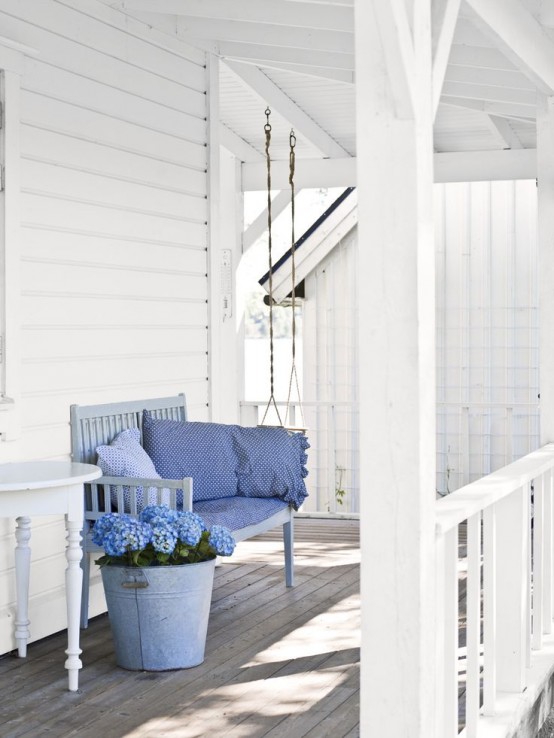a delicate vintage Scandi porch with a swing, a light blue sofa with pillows, a side table and some blooms in a bucket