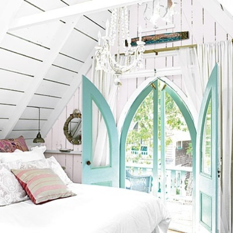 a white attic bedroom with a bed with neutral bedding, a white chandelier and turquoise framed glass doors that add color to the space and make it feel magical