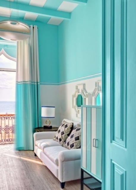 a bright turquoise living room with white paneling and neutral seating furniture, a striped wooden ceiling and color block turquoise curtains and turquoise doors