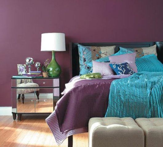 a deep purple bedroom with a black bed, colorful bedding including turquoise pillows and a blanket, a mirror nightstand, a table lamp and some books