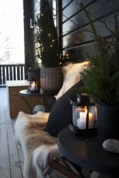 a winter terrace done with wooden furniture, faux fur, black metal side tables and potted Christmas trees with lights and lanterns on them
