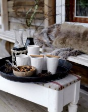 a relaxed Scandinavian terrace with a white bench and faux fur, with an ottoman with a tray, pinecones, hot drinks and cinnamon