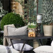 a small winter terrace with a folding table and chairs, neutral pillows, potted plants, greenery and a candle in a tall candleholder