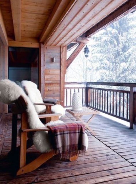 a winter terrace with a wooden deck, wooden chairs with faux fur and blankets and a wooden table with a vase plus a view of snowy forest